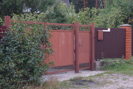 brown metal gate and a closed door on a fence wall on a rural street in green grass and bushes