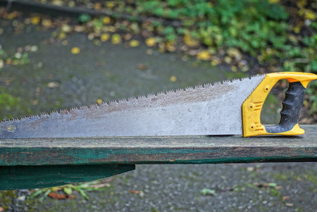 one gray old metal saw in rust with a yellow black plastic handle stands on a wooden table on the streetの写真素材