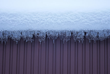 part of a brown metal wall under white snow with icicles on a winter street