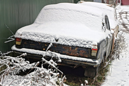 one old black rusty retro car under white snow stands on a winter street near a green fenceの写真素材