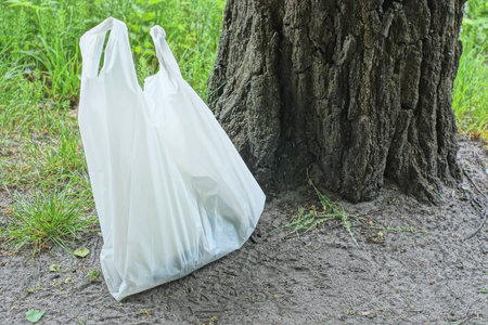 one white plastic bag with garbage stands on gray ground near green grass and a tree outdoors in natureの写真素材