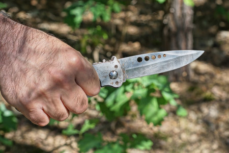 hand holds a gray steel hunting knife against the background of green plants on the street in nature
