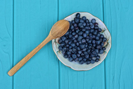 ripe fresh blueberries in a white plate with a brown spoon on a blue wooden tableの写真素材