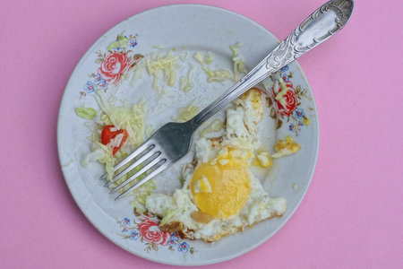 one white plate with a piece of yellow half-eaten yellow scrambled eggs with a metal gray fork on a...