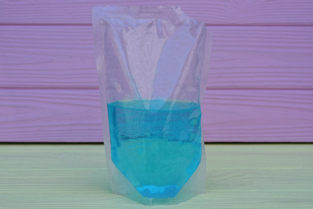 one white transparent plastic bag with blue liquid stands on a yellow wooden table against a pink...