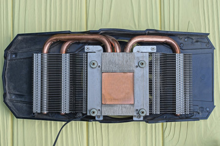 one black disassembled video card with a gray metal radiator and copper brown cooling pipes lies on a yellow wooden tableの写真素材