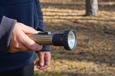 the hand of a man in blue clothes holds a metal electric flashlight on the street on a brown...