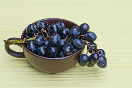 fresh blue ripe grapes on a branch in a brown ceramic cup stands on a yellow wooden table
