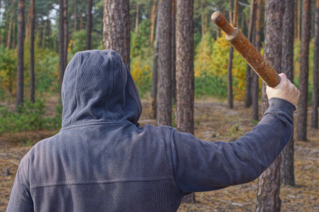 an aggressive man in blue clothes and a hood swings a brown stick with a bat over his head on the street among the trees in the forestの写真素材