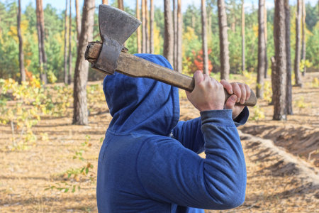 one male criminal in blue clothes and a hood swings hands a big ax aggressively on the street among the treesの写真素材