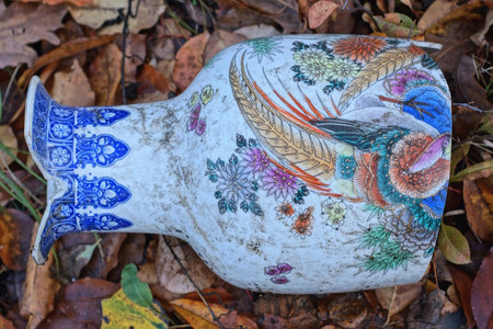 one white dirty broken ceramic Chinese vase with a colorful pattern lies on the ground and fallen leaves on the streetの写真素材