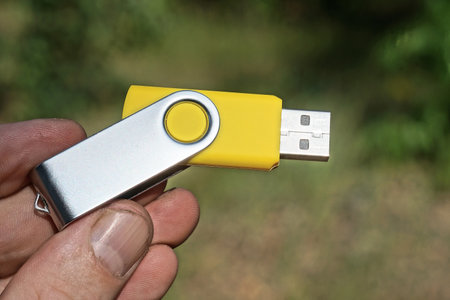 fingers on the hand hold one gray yellow small compact flash drive outdoors on a green background