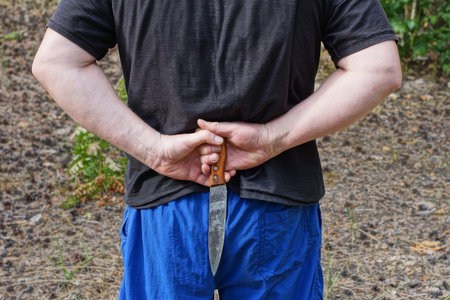 hands of a male in black clothes and a gray long knife behind his back on the street