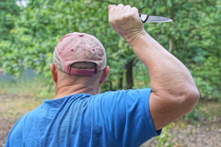 one aggressive man in blue clothes and red cap waves a knife in his hand over his head on the streetの写真素材