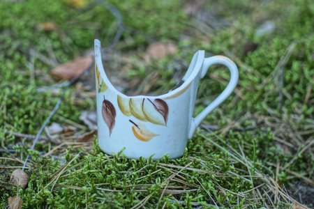 one white old broken ceramic cup with colorful pattern stands on green moss on summer street in natureの写真素材
