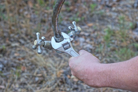 worker's hand with gray adjustable wrench unscrews nut on iron old water tap on summer streetの写真素材