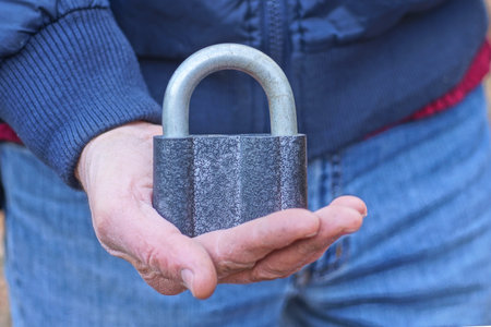 one old big gray iron padlock hanging on a finger on a hand in the streetの写真素材