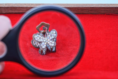 one black magnifying glass in jeweler's hand magnifies small old copper jewelry pin with colored stones on red tableの写真素材