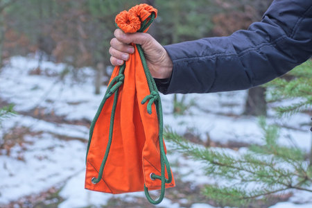 one hand of a man in black clothes holds an orange bag with a green strap on a winter street against...