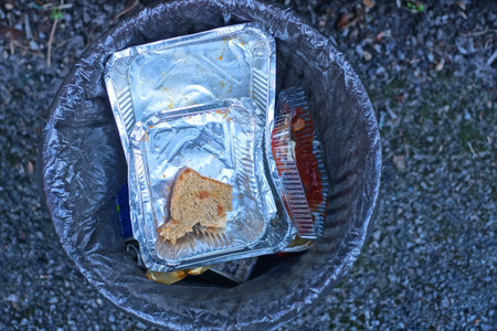 one trash can with gray plastic bag inside and garbage from foil dishes and pieces of food on gray floor on summer streetの写真素材