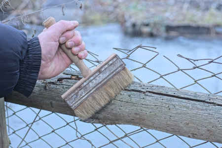 one hand of worker holds gray brown big paint brush painting wooden board on old metal mesh fence on autumn streetの写真素材