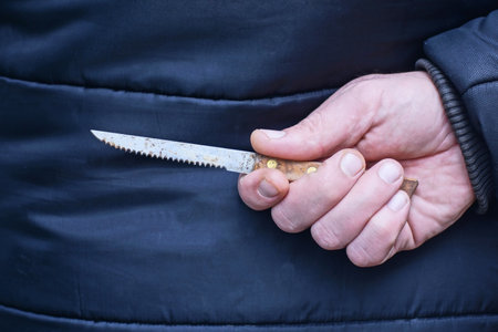 hand of a male in black clothes and a gray knife behind his back on the streetの写真素材