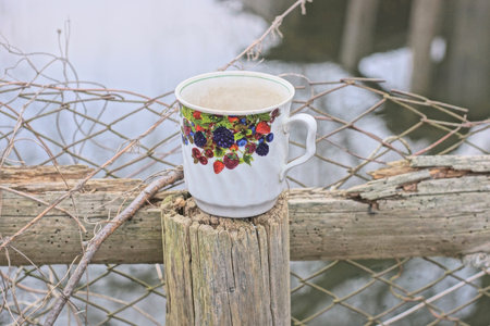 one white ceramic cup with colorful pattern stands on gray wooden post of metal mesh fence on autumn...