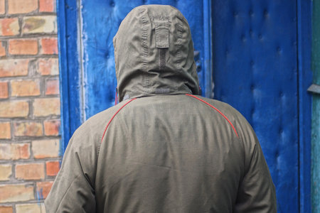 one man stands with his back in a green jacket with a hood stands on an autumn street against a blue wallの写真素材