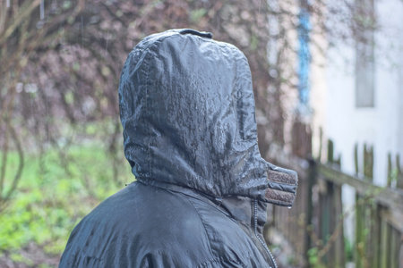 a man stands in a gray green wet jacket with a hood on his head in the rain on an autumn street