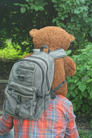 one man in colorful clothes holding big brown toy bear with gray backpack on green vegetation backgroundの写真素材