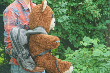 one man in colorful clothes holding big brown toy bear with gray backpack on green vegetation background on summer streetの写真素材