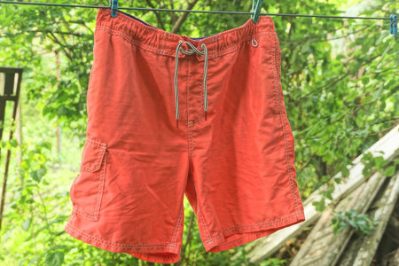 one wet shorts made of red fabric hanging on a wire against the background of green vegetation on a summer streetの写真素材