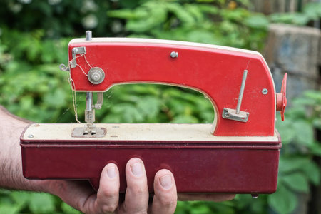 hand holds one old red white toy sewing machine near green vegetation on a summer street
