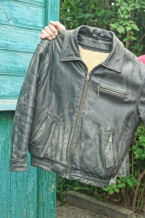 man's hands holding old black leather jacket on summer street against green wall