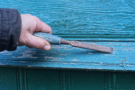 hand holds brown old rusty iron chisel in green wood plank wallの写真素材