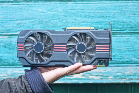 A hand holds one large black video card with two fans against a green wall on a summer streetの写真素材