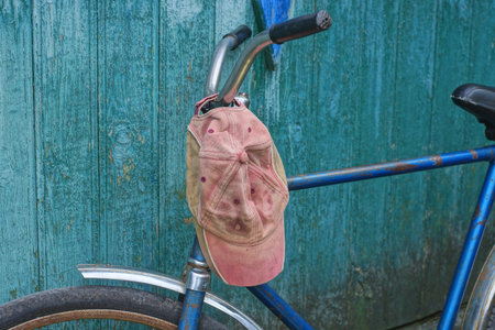 One red old cap hangs on the handlebars of a bicycle near a green wall on a summer streetの写真素材