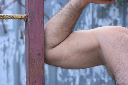 one arm of a male athlete bend in tension at a brown metal pole outdoors