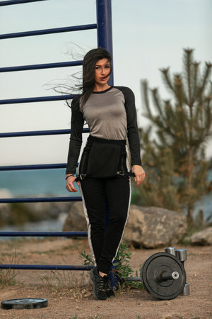 young woman stands by a gym apparatus in an outdoor setting. She wears casual athletic attire and...