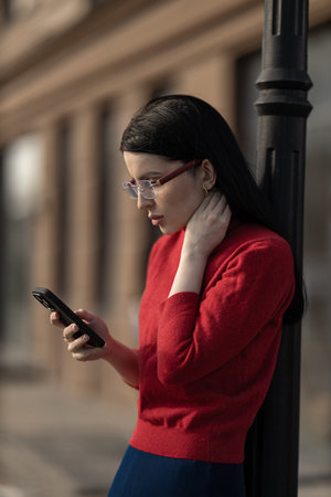 beautiful girl wearing glasses stands outdoors, leaning against a street lamp, intently looking at her phone in a warm, sunny urban environment She wears a stylish outfit that complements her lookの写真素材