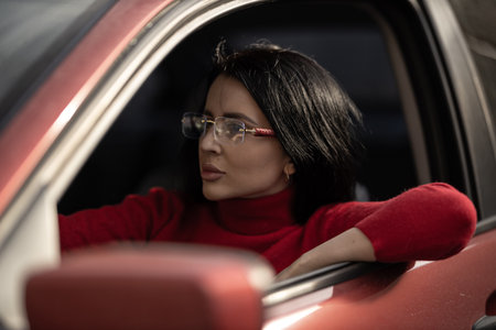 beautiful girl wearing stylish glasses leans out of a car window, dressed in a cozy red sweater She...