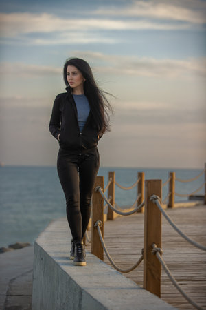 woman stands pensively on a coastal boardwalk, gazing at the ocean as the sun sets, casting a warm glow across the calm water and skyの写真素材