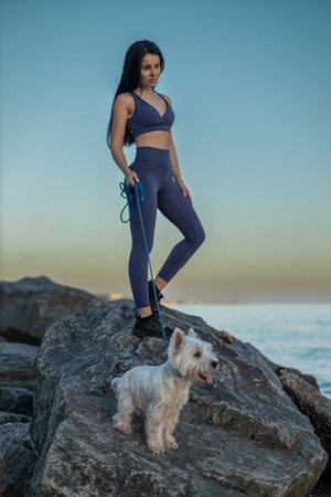beautiful brunette woman wearing fitness attire stands on a rock by the beach She enjoys a serene sunset stroll with her white dog beside her, showcasing her smooth skin after epilationの写真素材