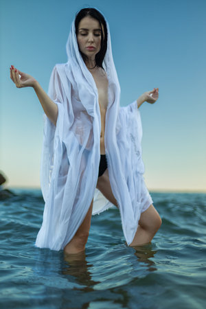 beautiful brunette woman stands gracefully in the tranquil sea wearing a flowing white garment The sun sets in the background, casting a serene glow around herの写真素材