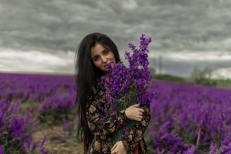 beautiful brunette stands in a vibrant field of delphiniums, holding a bouquet of purple flowers Dark clouds loom above as she smiles, capturing the essence of natures beautyの写真素材