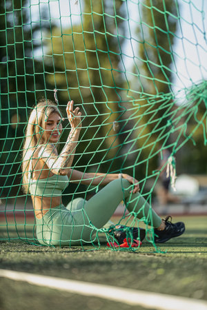 young woman in green athletic wear strikes playful pose while sitting against sports net warm sunlight highlights her outfit and vibrant surroundingsの写真素材