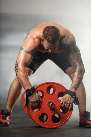 man with tattoos in gym leans forward to hang red weight on barbell positioned on the floor focused...