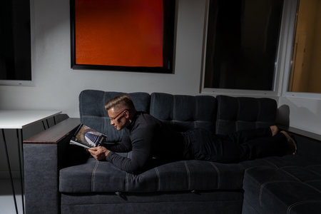 athletic man with glasses enjoys reading on a comfortable sofa in a softly lit room at night. The atmosphere is relaxed and peaceful perfect for unwinding with a good book.の写真素材