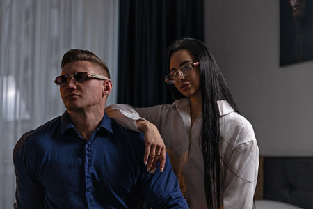Man in a blue shirt and woman in a white shirt stand close together both wearing glasses. They are...
