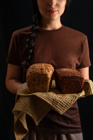 brunette girl in a brown T shirt stands indoors holding two loaves of bread on a golden cloth. The...